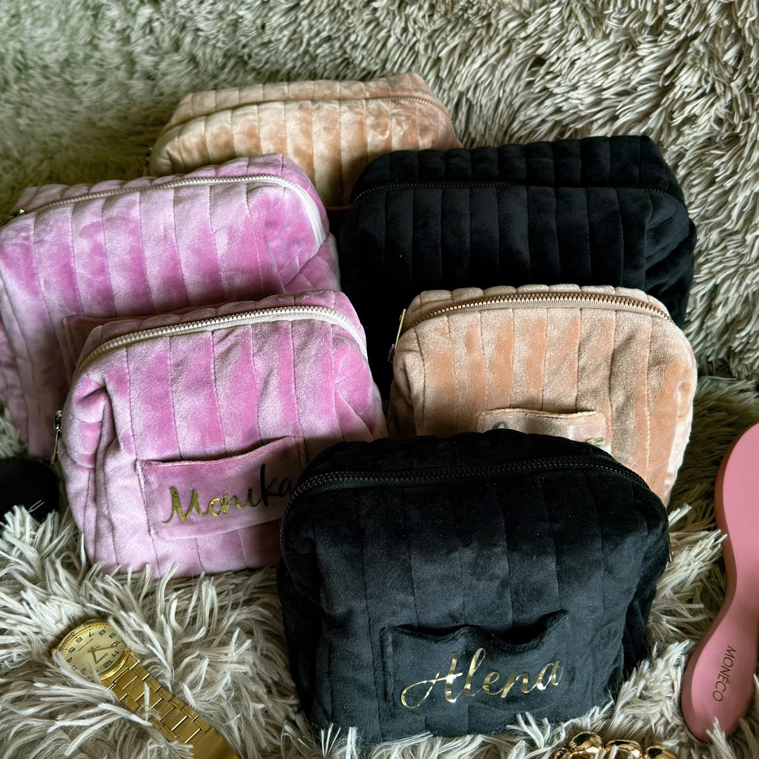 Personalized velvet cosmetic bags in pink, beige, and black on plush surface with watch and brush